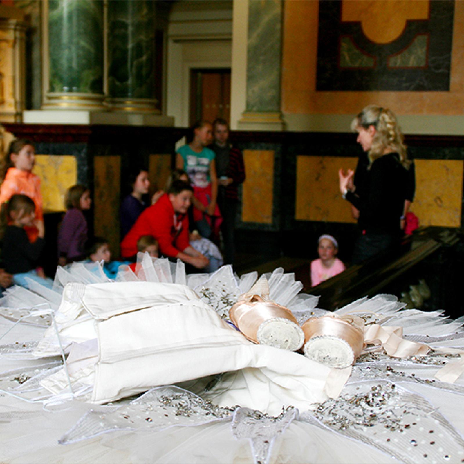 Ein Paar Spitzenschuhe und ein verzierter Tutu liegen im Vordergrund, während im Hintergrund eine Gruppe von Kindern einer Führerin in einem prachtvollen historischen Gebäude zuhört.