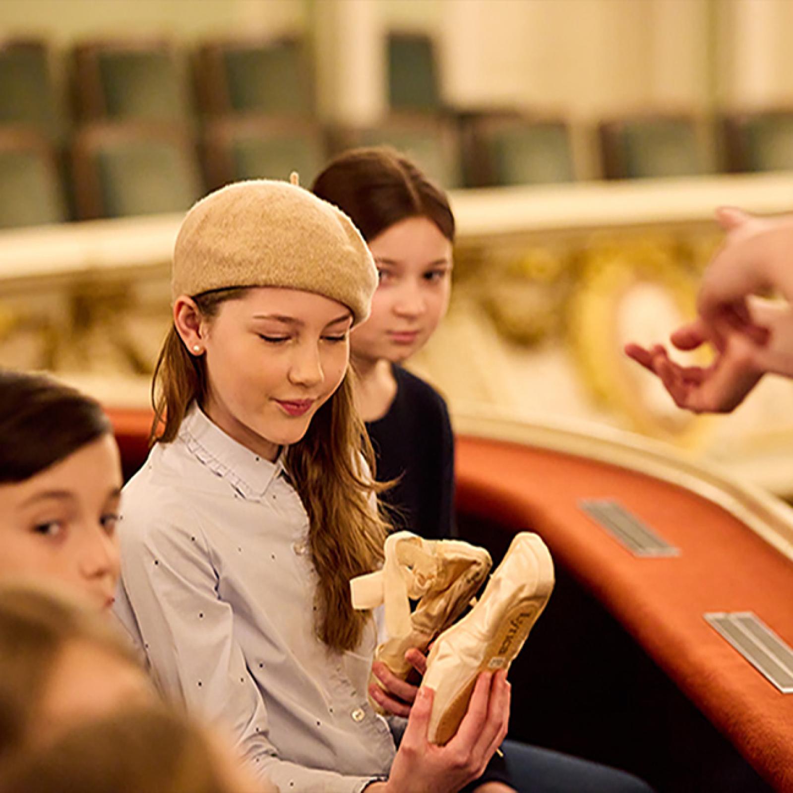 Ein Mädchen mit beigefarbener Baskenmütze betrachtet ein Paar Ballettspitzenschuhe, während andere Kinder neben ihr in einer kunstvoll gestalteten Opernloge sitzen.