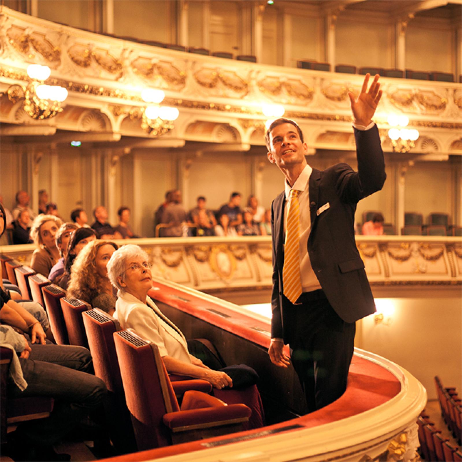 Ein Guide im Anzug steht in einer verzierten Opernloge und deutet in den Zuschauerraum, während die Besucher aufmerksam sitzen und dem Rundgang im prächtigen Opernhaus folgen.