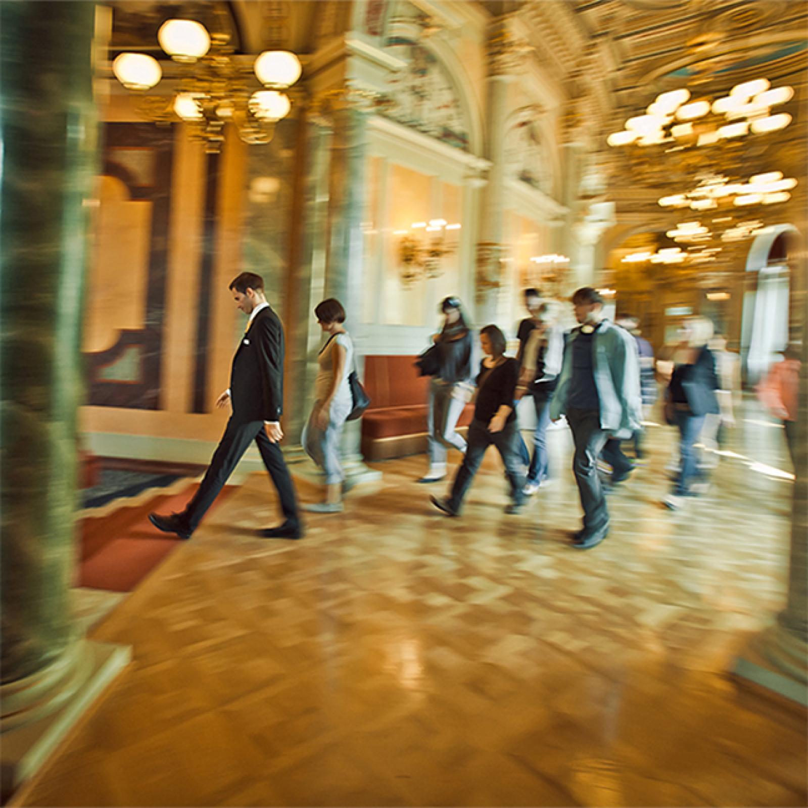 Eine Führungsgruppe geht durch einen prunkvollen Flur mit Marmorsäulen und Kronleuchtern, angeführt von einem Guide im Anzug; die Bewegungsunschärfe verstärkt den Eindruck des Voranschreitens durch das historische Interieur.