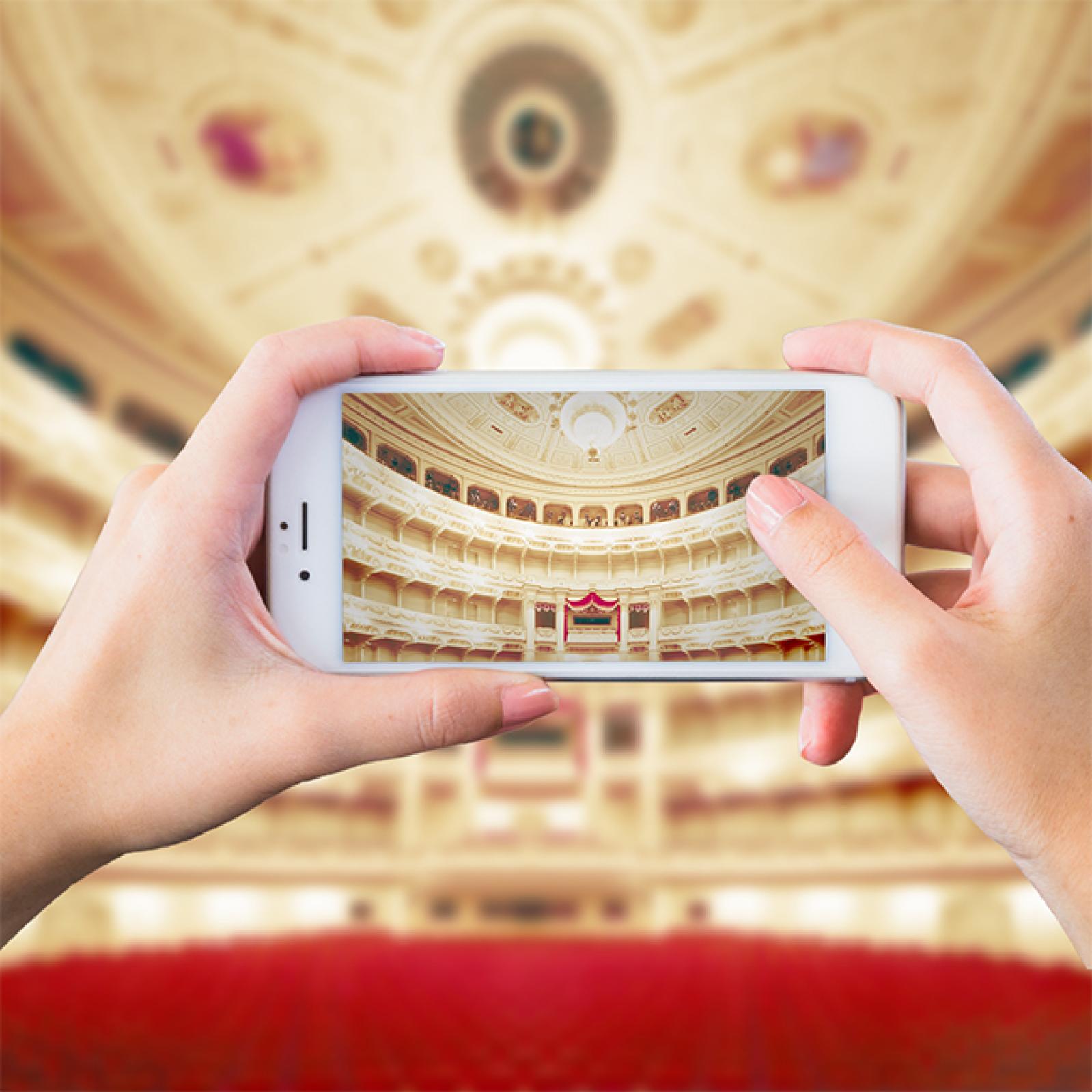 Hände halten ein Smartphone, das den prunkvollen Zuschauerraum eines Opernhauses mit roten Sitzreihen, Logen und reich verzierter Decke fotografiert.
