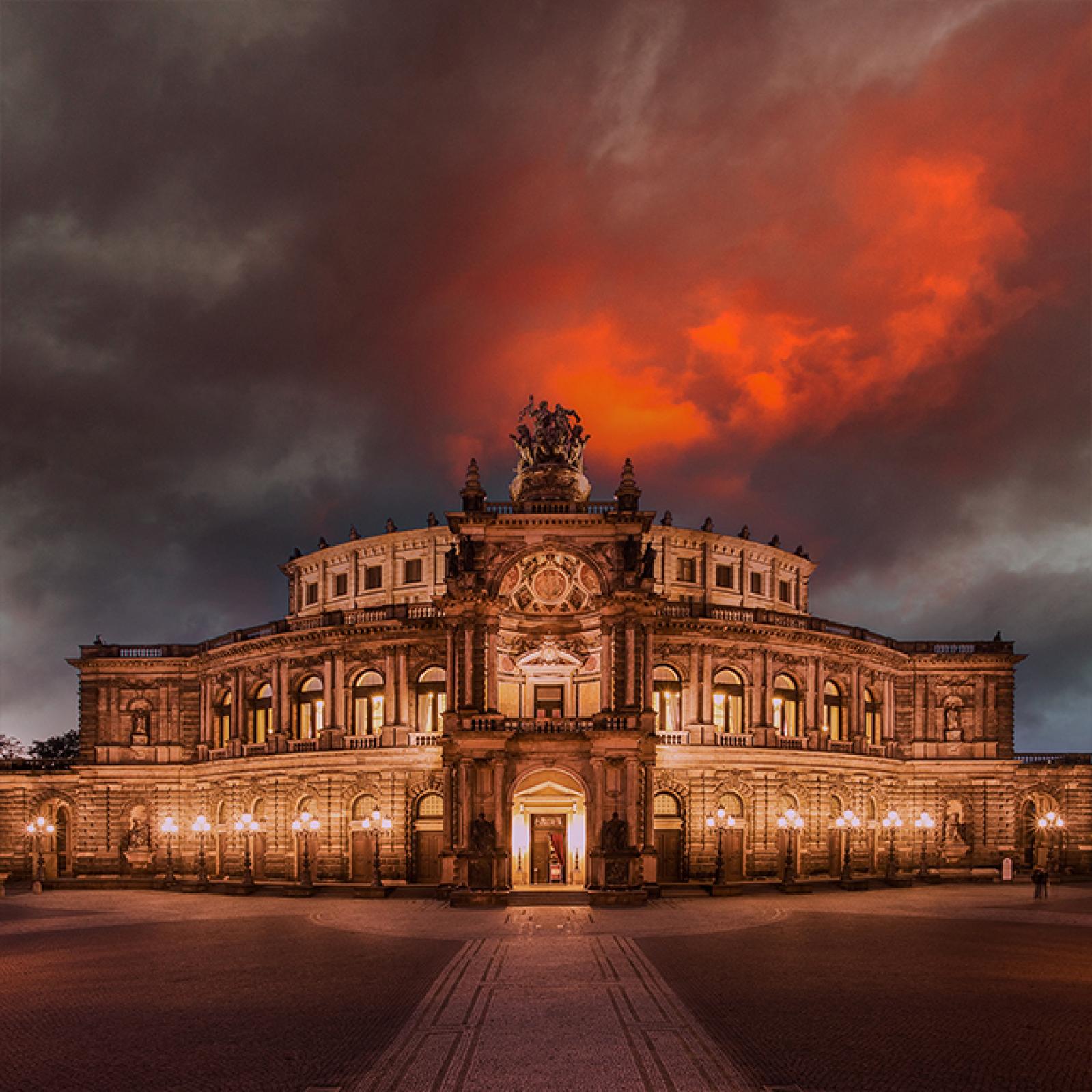 Die Dresdner Semperoper erleuchtet in der Dämmerung, darüber dramatische dunkle Wolken, die in feurigem Orange glühen und eine geheimnisvolle, stimmungsvolle Szenerie erzeugen.