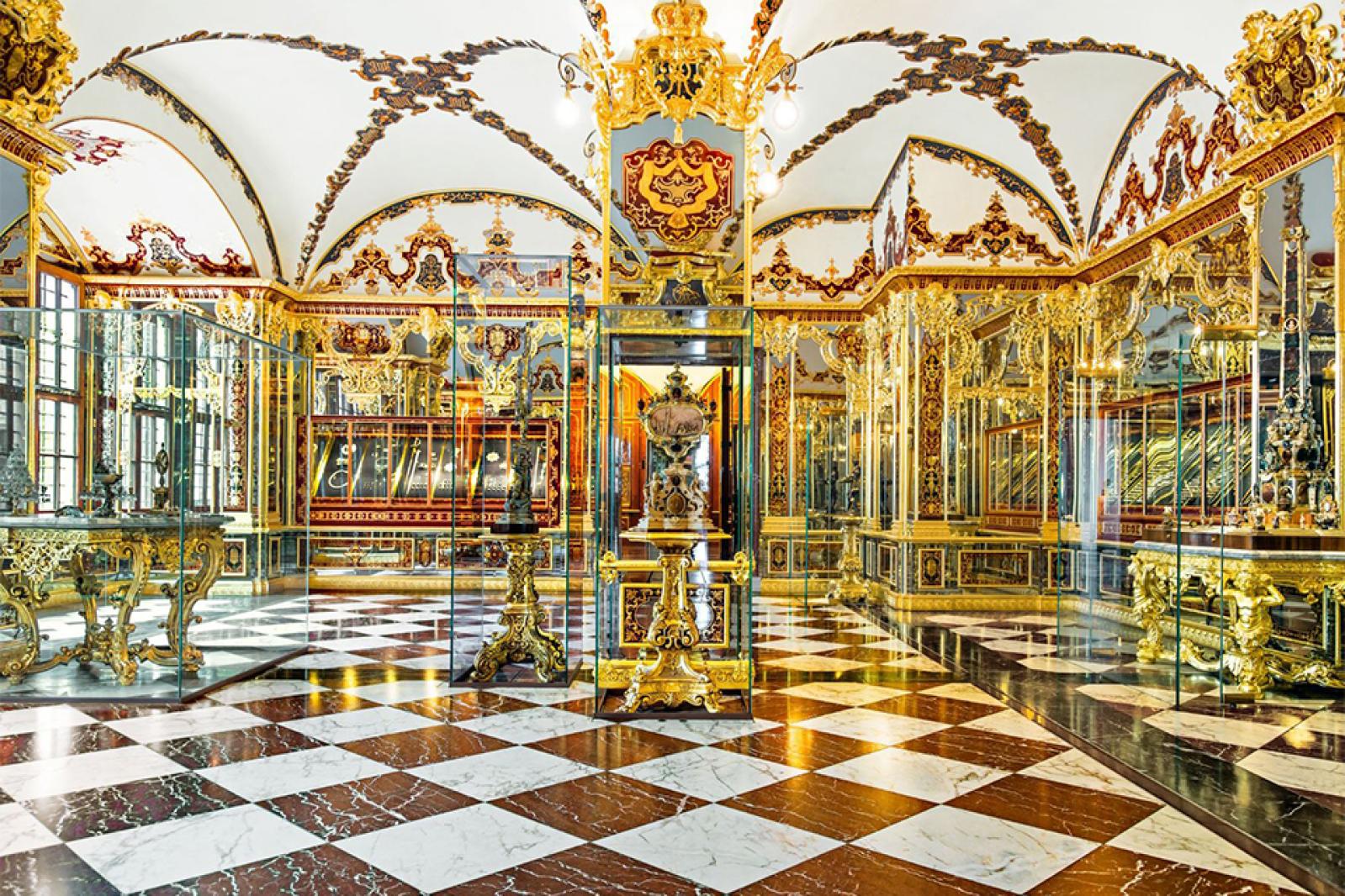Ein prunkvoller Museumsraum mit vergoldeten Vitrinen, verspiegelten Wänden, reich verzierten barocken Dekorationen und einem schwarz-weiß karierten Marmorboden zur Präsentation historischer Kostbarkeiten.