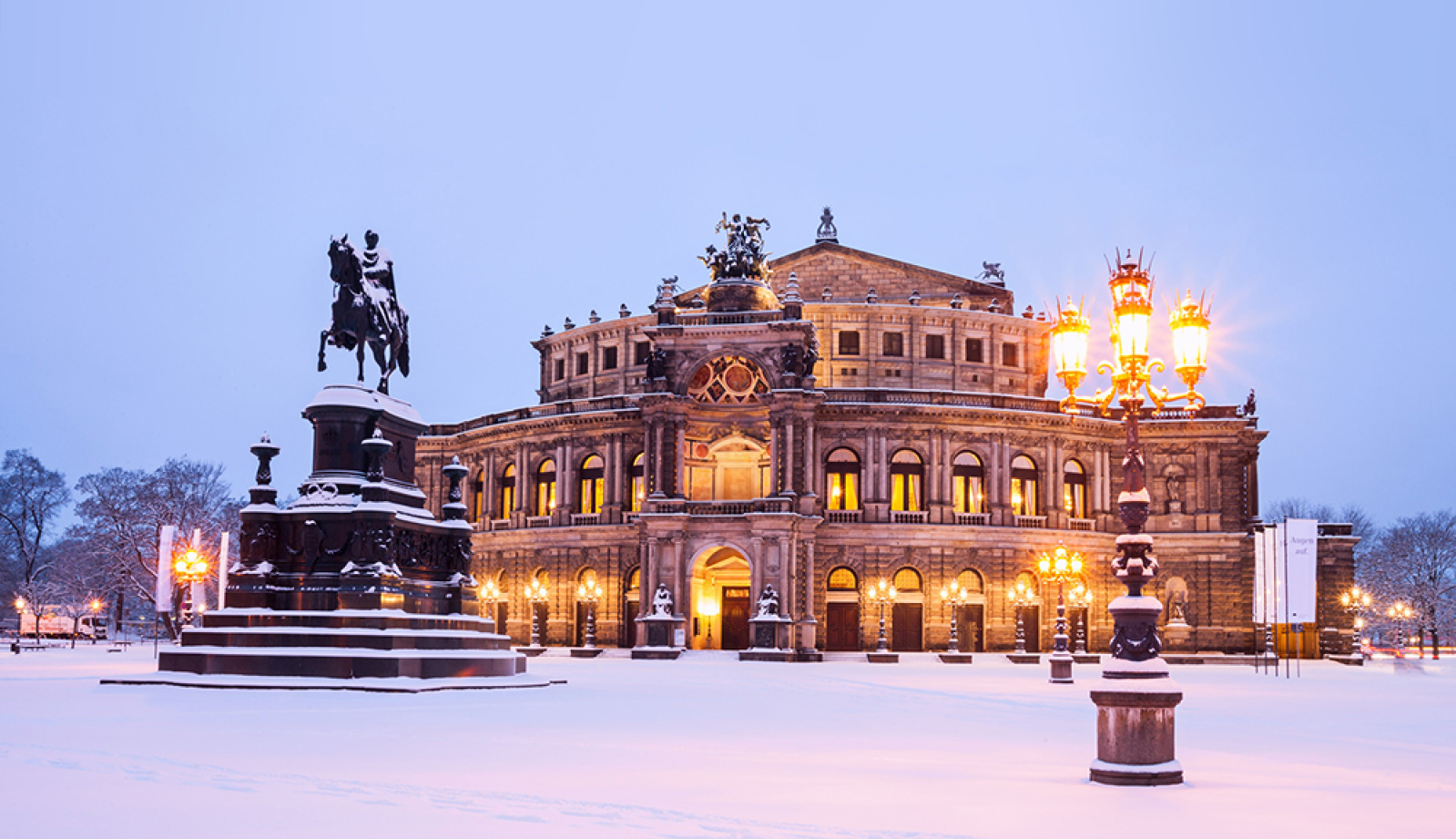 Die Semperoper in Dresden erstrahlt an einem winterlichen Abend im Schnee, mit leuchtenden Laternen und dem Reiterstandbild im Vordergrund.