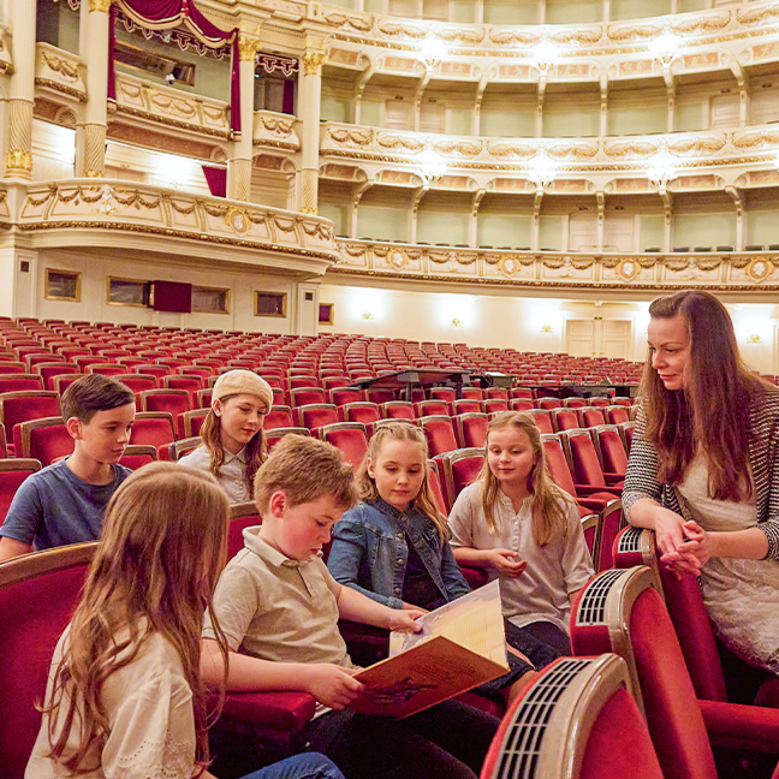Eine Gruppe von Kindern sitzt gemeinsam in den roten Samtsesseln des Zuschauerraums der Semperoper, betrachtet ein aufgeschlagenes Buch und wird dabei von einer Führerin im Rahmen einer Familienführung begleitet.