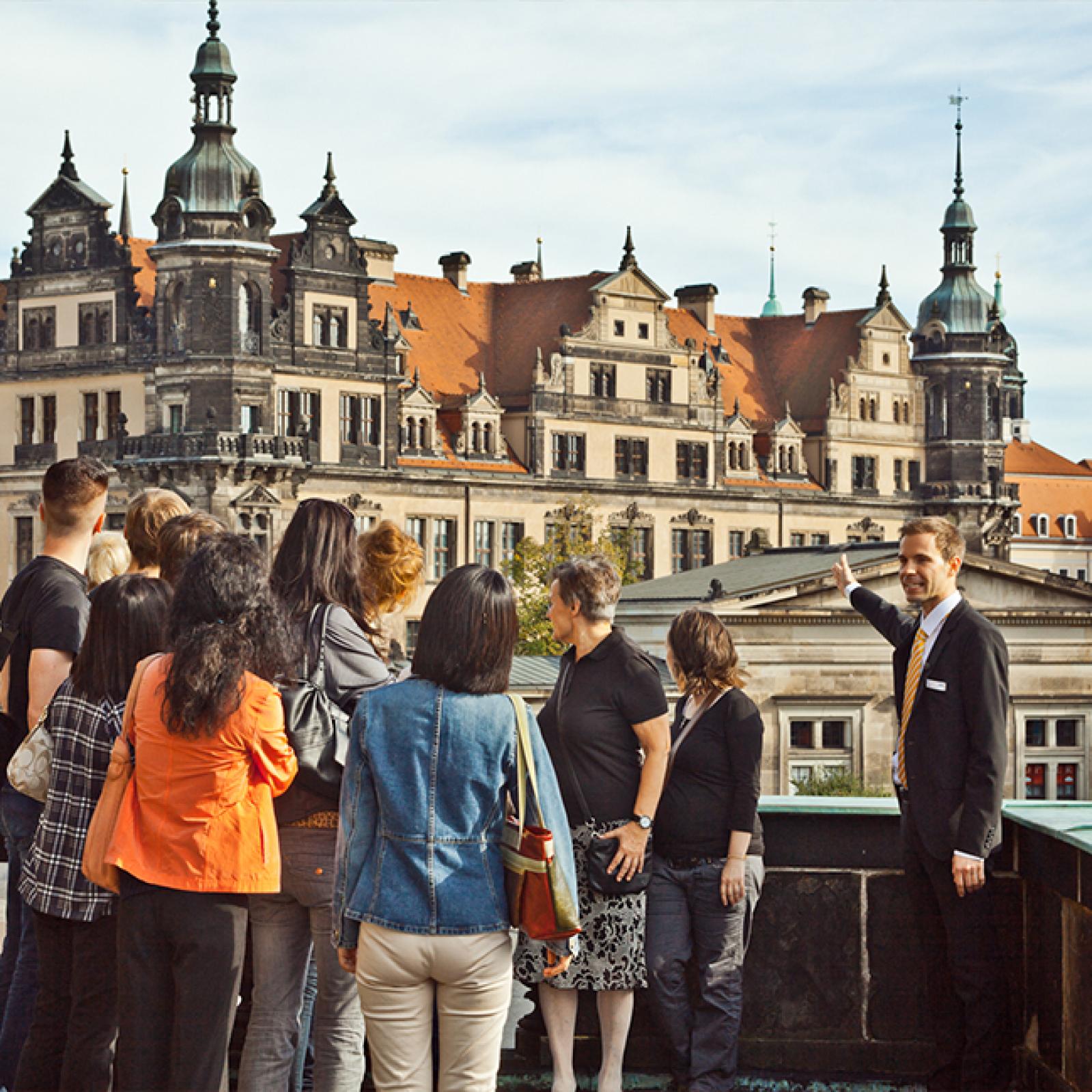 Ein Tourguide steht mit einer Besuchergruppe auf einer Terrasse und zeigt auf ein historisches Barockgebäude mit Türmen und rotem Dach in einer europäischen Altstadt.