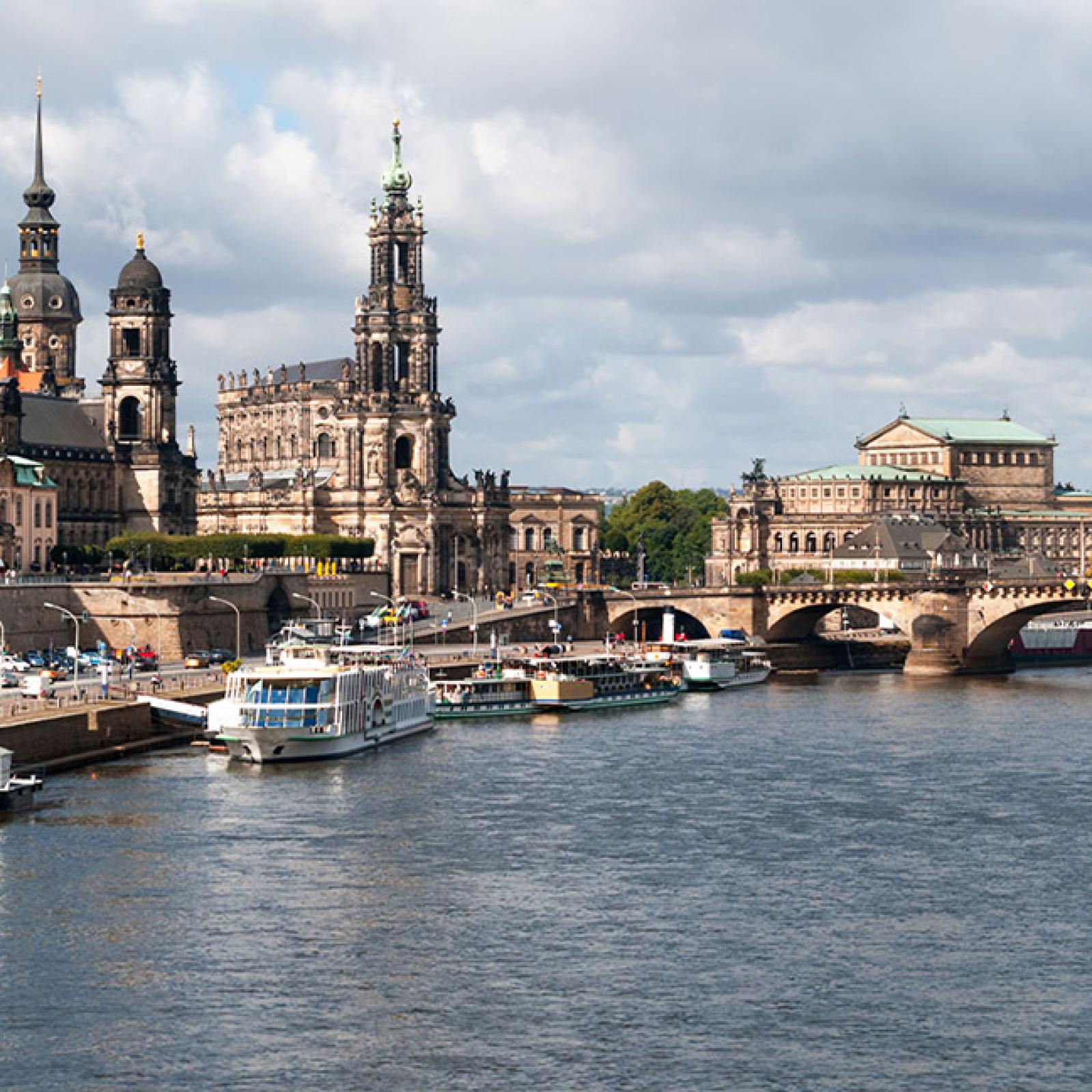 Blick entlang der Elbe in Dresden mit historischen Gebäuden, Brücken und Ausflugsschiffen, darunter die Silhouette der Altstadt bei leicht bewölktem Himmel.