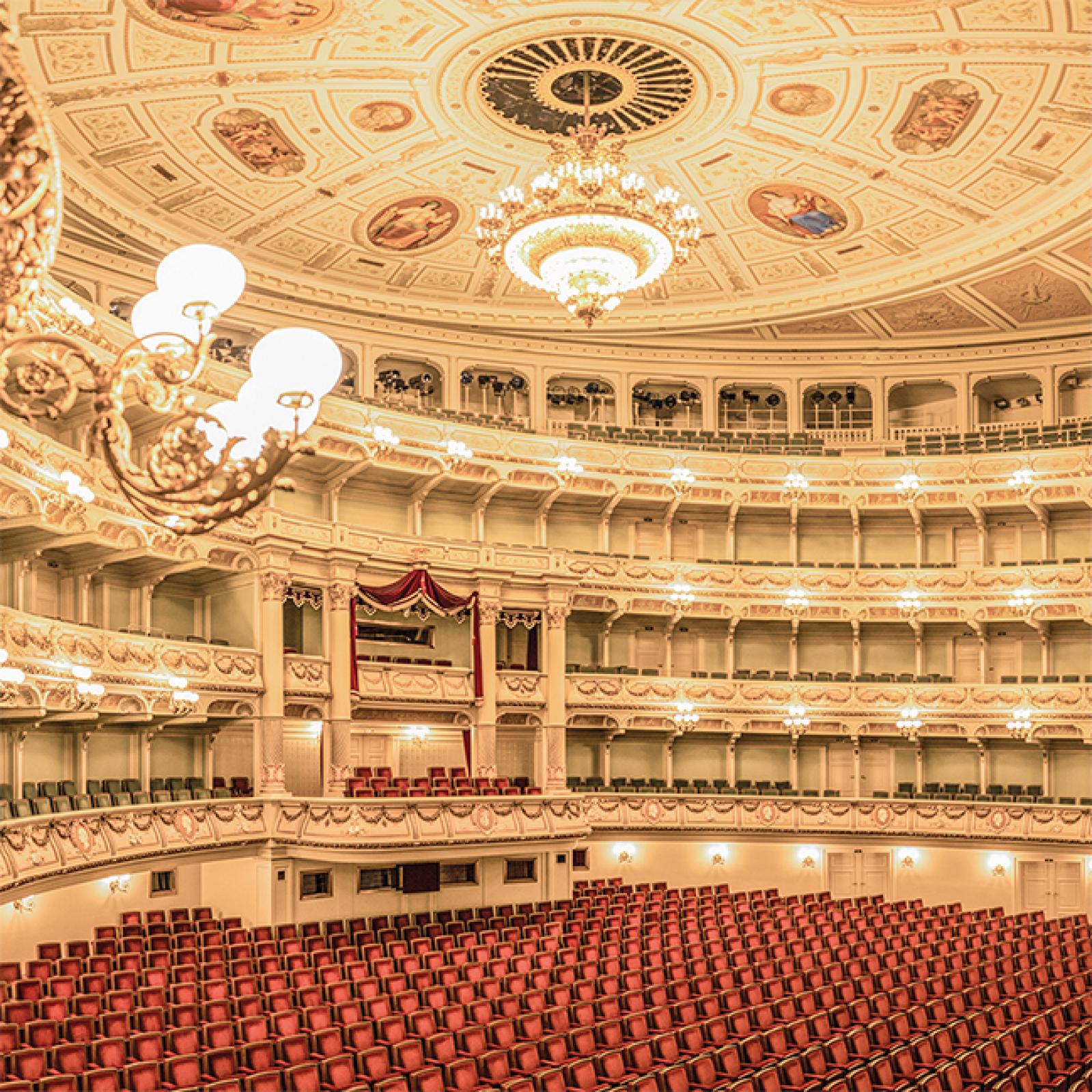 Der prachtvolle Zuschauerraum eines Opernhauses mit roten Sitzreihen, mehreren Balkonetagen, funkelnden Kronleuchtern und einer kunstvoll bemalten Decke.