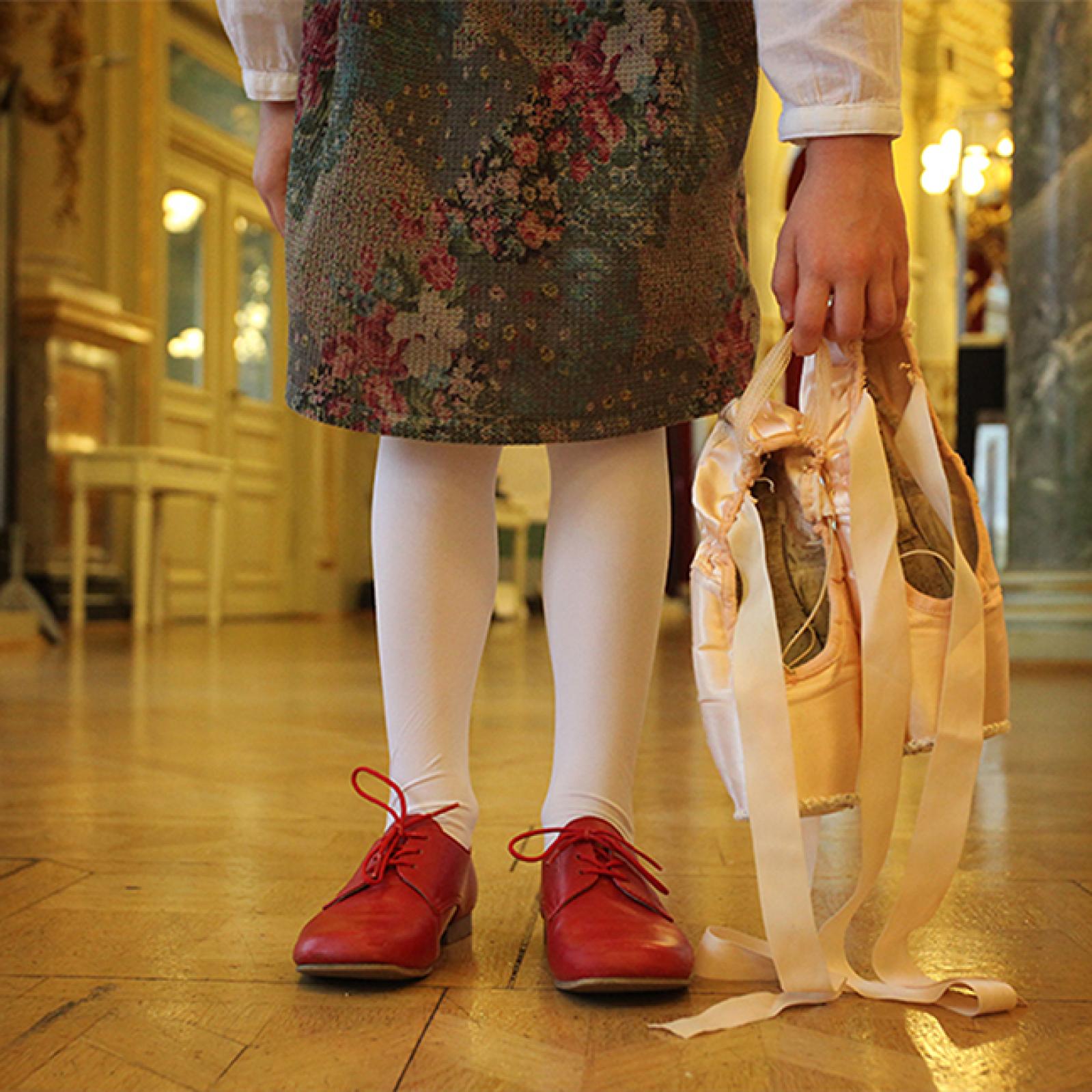 Ein junges Mädchen steht in einem prachtvollen Saal, trägt weiße Strumpfhosen und rote Schuhe und hält ein Paar satinierte Ballettspitzenschuhe an den Bändern in der Hand.