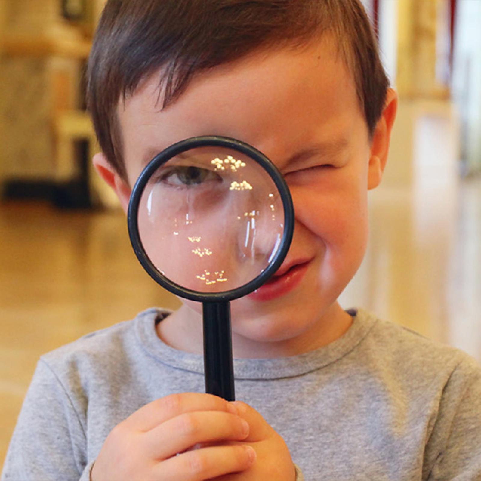 Ein kleiner Junge hält sich spielerisch eine Lupe vor ein Auge und blinzelt mit dem anderen, in einem hell beleuchteten Raum.