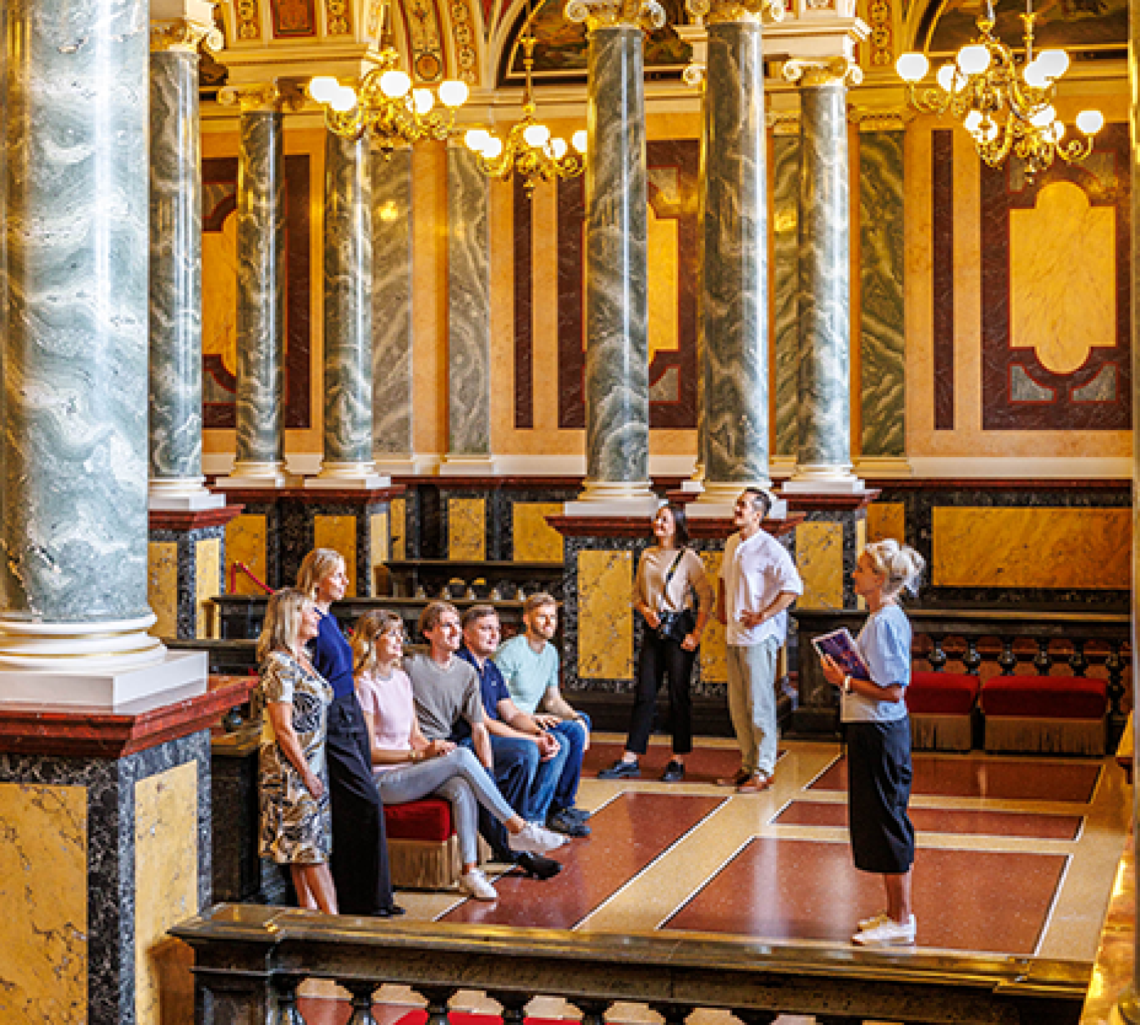 Eine geführte Gruppe innerhalb eines prunkvollen Interieurs mit hohen grünen Marmorsäulen, verzierten Bögen, Kronleuchtern und kunstvoll bemalten Decken in einem historischen Opernhaus.