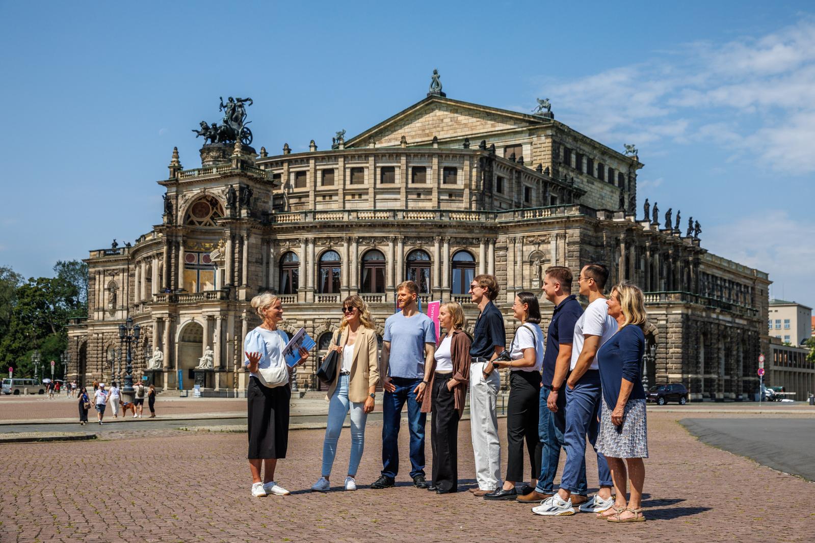 Ein Tourguide steht mit einer Besuchergruppe auf einer Terrasse und zeigt auf ein historisches Barockgebäude mit Türmen und rotem Dach in einer europäischen Altstadt.