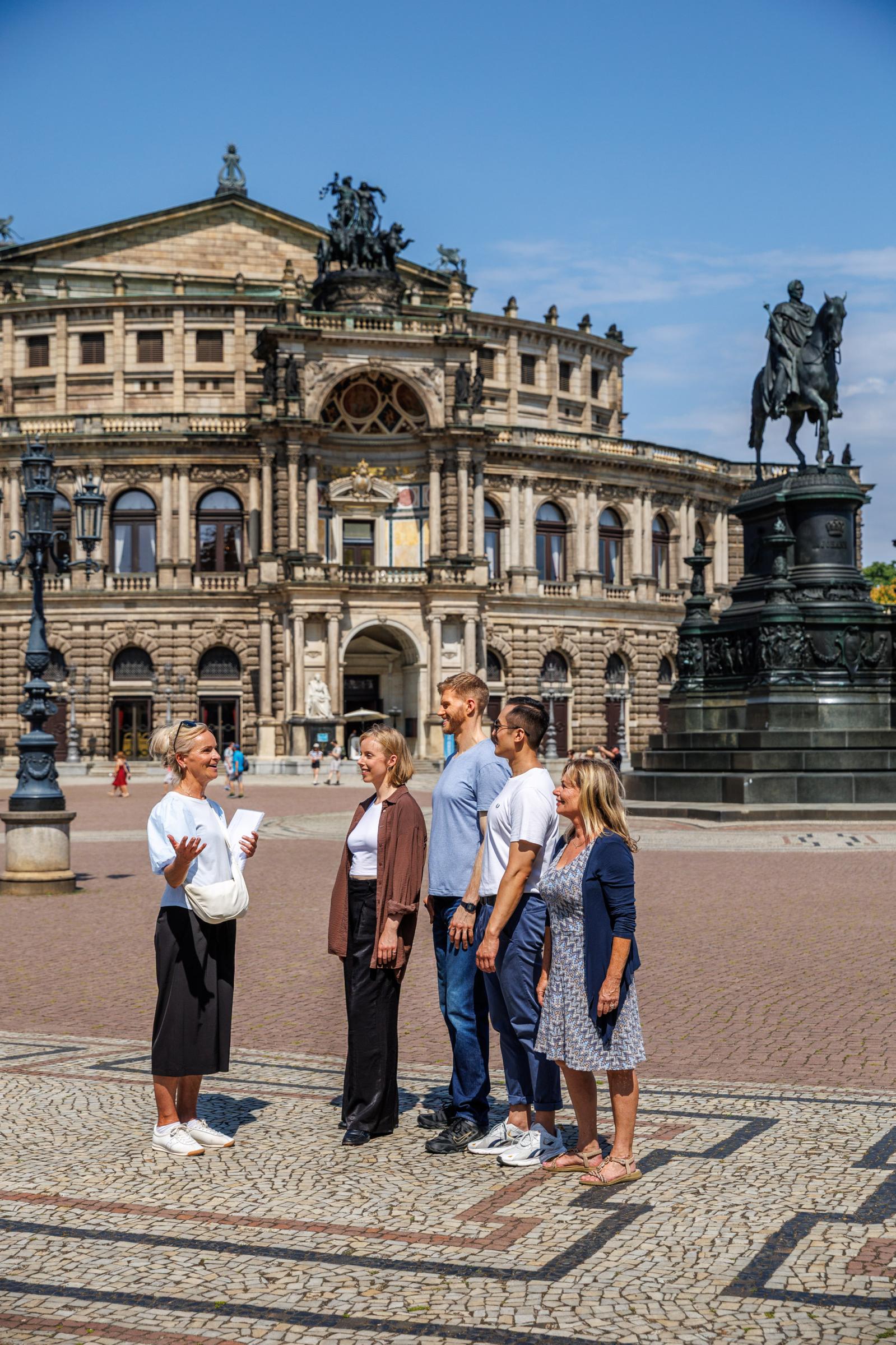 Ein Tourguide steht mit einer Besuchergruppe auf einer Terrasse und zeigt auf ein historisches Barockgebäude mit Türmen und rotem Dach in einer europäischen Altstadt.