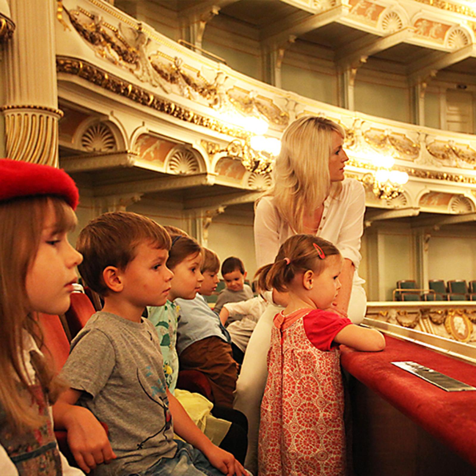 Eine Gruppe kleiner Kinder sitzt in einer kunstvoll gestalteten Opernloge und hört aufmerksam zu, begleitet von einer Erwachsenen, die sich neben ihnen nach vorn lehnt.