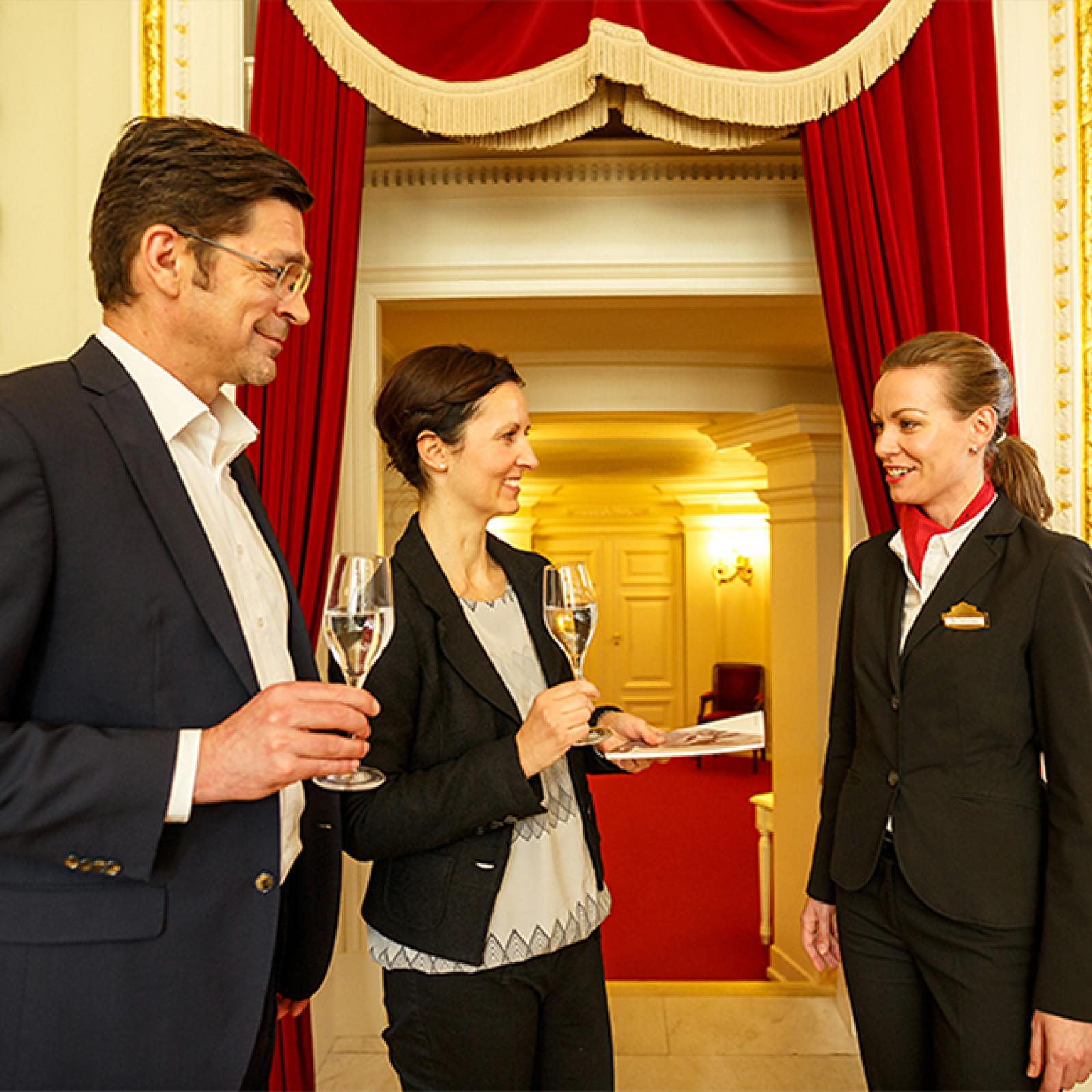 Ein elegant gekleidetes Paar mit Gläsern Sekt unterhält sich mit einer Mitarbeiterin des Opernhauses, stehend vor roten Samtvorhängen in einem repräsentativen Innenraum.