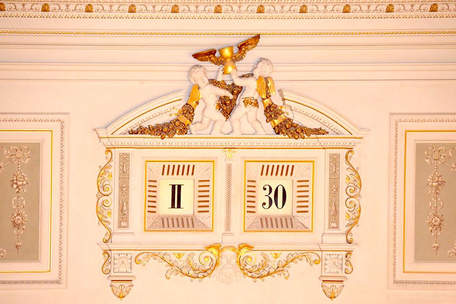 Eine kunstvoll gestaltete historische Uhr im Inneren der Semperoper mit goldenen Verzierungen, zwei Putten unter einem Adler sowie römischen Ziffern und der Zahl 30.