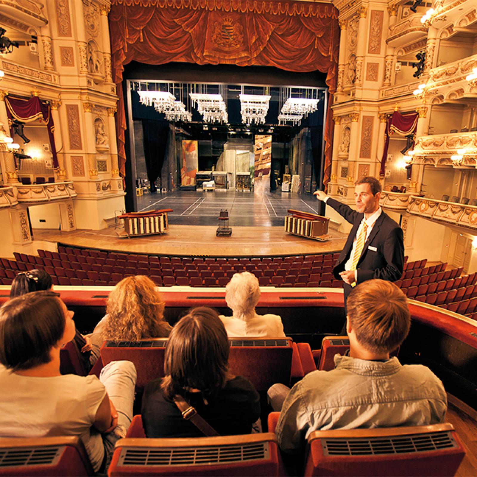 Ein Guide steht mit Besuchern in einer Opernloge und zeigt auf die Bühne, auf der Bühnenelemente und Beleuchtungsgerüste zu sehen sind – ein Blick hinter die Kulissen in einem prachtvollen Opernhaus.