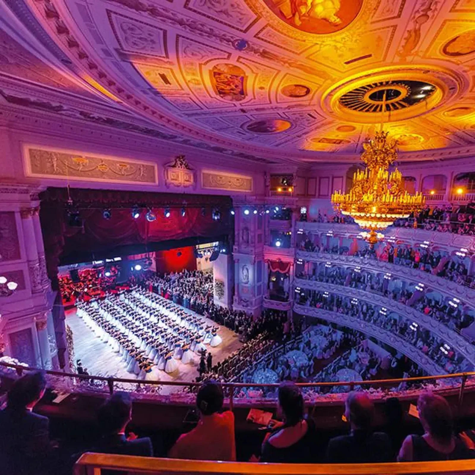 Großzügiger Blick in den Zuschauerraum der Semperoper während des SemperOpernballs, mit Orchester im Parkett, elegant gekleideten Gästen auf mehreren Rängen und einem leuchtenden Kronleuchter unter farbiger Lichtinszenierung.