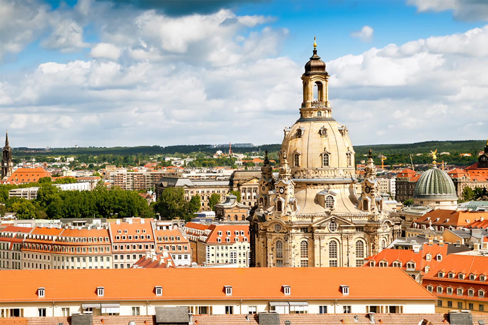 Panoramablick über die Dresdner Altstadt mit der Frauenkirche und zahlreichen rot gedeckten Dächern unter blauem Himmel.
